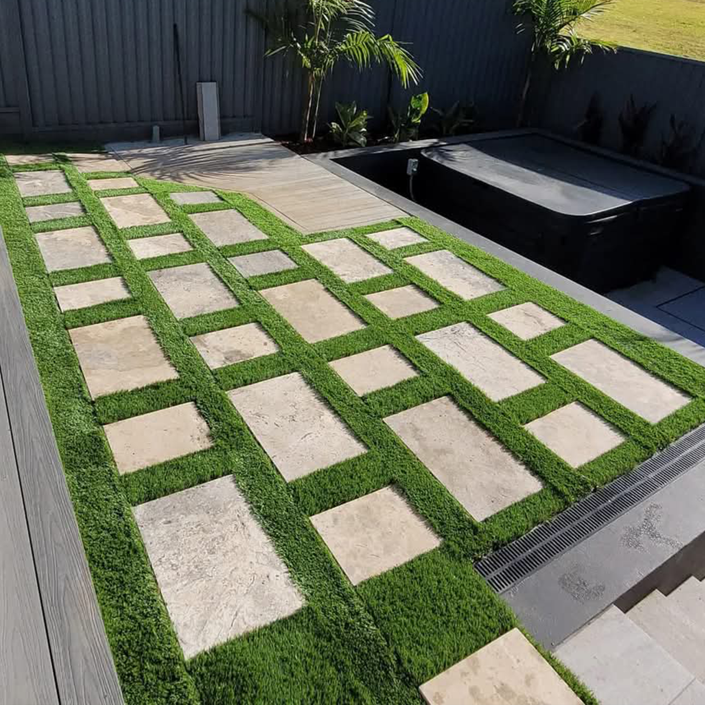 Modern Sydney patio featuring a geometric stone paver grid with grass strips, timber deck and tropical plants.